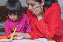 A mother and her young female child seated at a desk and leaning in together to look at a children’s workbook.