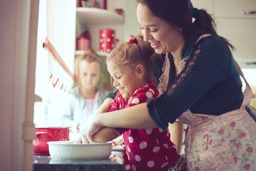 An adult standing behind a young child smiling while mixing ingredients in a bowl together. Another slightly older child watches solemnly.